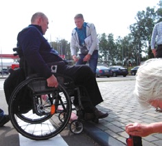 на фотографии изображен человек сидящий на инвалидной коляске перед бордюром и эксперты замеряющие нормативы