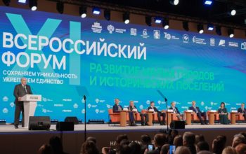 На фотографии запечатлено выступление спикера за трибуной на сцене Всероссийского форума «Развитие малых городов и исторических поселений», где на фоне масштабного синего экрана с логотипами и лозунгами мероприятия на креслах сидят другие участники президиума, а перед ними в зале находится аудитория.