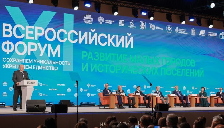 На фотографии запечатлено выступление спикера за трибуной на сцене Всероссийского форума «Развитие малых городов и исторических поселений», где на фоне масштабного синего экрана с логотипами и лозунгами мероприятия на креслах сидят другие участники президиума, а перед ними в зале находится аудитория.