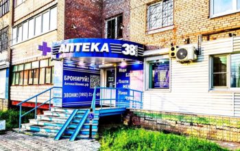 На фотографии запечатлен вход в «Аптеку 38 плюс», расположенную на первом этаже кирпичного жилого дома и оборудованную крыльцом со ступенями, металлическими рельсовыми аппарелями для колясок, синей вывеской и рекламными баннерами со слоганами и QR-кодами.