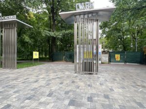 На фотографии запечатлена вымощенная плиткой входная группа в парк с высокими металлическими пилонами-стелами, на фоне зелёных деревьев и строительного ограждения. 