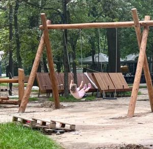 На фотографии запечатлен ребенок в розовой одежде, качающийся на цепных качелях с массивной деревянной А-образной опорой, на фоне деревянных шезлонгов, зеленых деревьев и лежащего на земле строительного поддона.