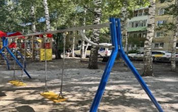 На фотографии запечатлена детская площадка с качелями на цепях во дворе жилого дома, где на заднем плане среди берёз припаркованы автомобили, включая машину скорой помощи.