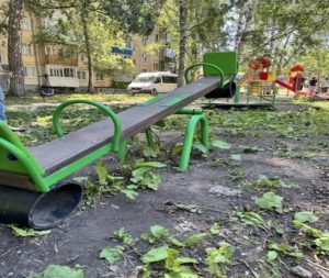 На фотографии запечатлена детская площадка во дворе жилого дома, где на переднем плане крупным планом показаны деревянно-металлические качели-балансир с зелёными поручнями, установленные на неровной земле с редкой травой на фоне деревьев, карусели и припаркованного автомобиля.