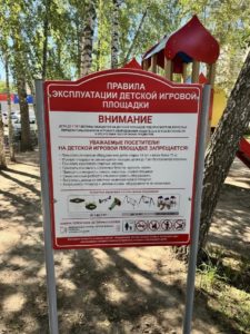 На фотографии крупным планом запечатлен установленный на детской площадке информационный стенд с красным заголовком «Правила эксплуатации детской игровой площадки», на котором подробно расписаны меры безопасности и запреты для посетителей.
