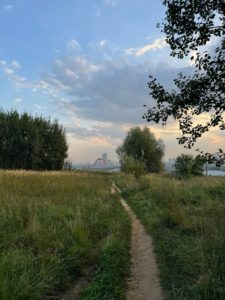 На фотографии запечатлена узкая грунтовая тропинка, петляющая среди высокой травы Крылатских холмов по направлению к виднеющемуся вдали Живописному мосту под предзакатным небом с мягкими облаками.