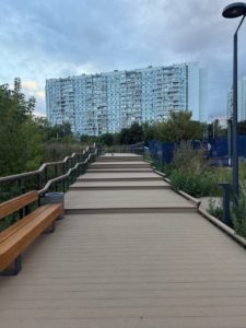 На фотографии запечатлена широкая деревянная экотропа с лавочкой и перилами, переходящая в ступенчатый подъем, на фоне многоэтажного жилого дома и пасмурного неба.
