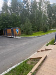 На фотографии запечатлен современный общественный туалет с пандусом, установленный возле асфальтированной дорожки и деревьев, вид на который открывается со стороны деревянного настила с пандусом-съездом.