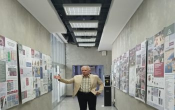 На фотографии запечатлен С.В. Чистый в светлом пиджаке и клетчатой рубашке, который жестом руки указывает на информационные плакаты и чертежи проектов благоустройства, развешанные вдоль стен длинного современного коридора со встроенным потолочным освещением.