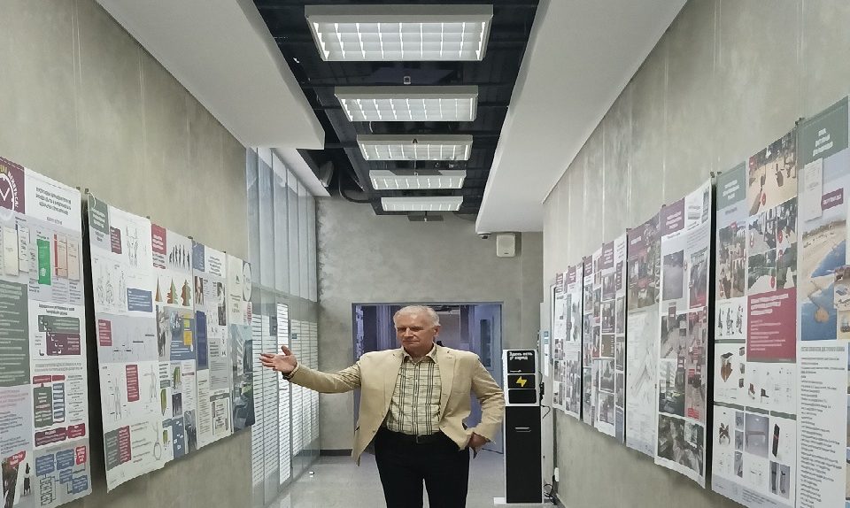 На фотографии запечатлен С.В. Чистый в светлом пиджаке и клетчатой рубашке, который жестом руки указывает на информационные плакаты и чертежи проектов благоустройства, развешанные вдоль стен длинного современного коридора со встроенным потолочным освещением.