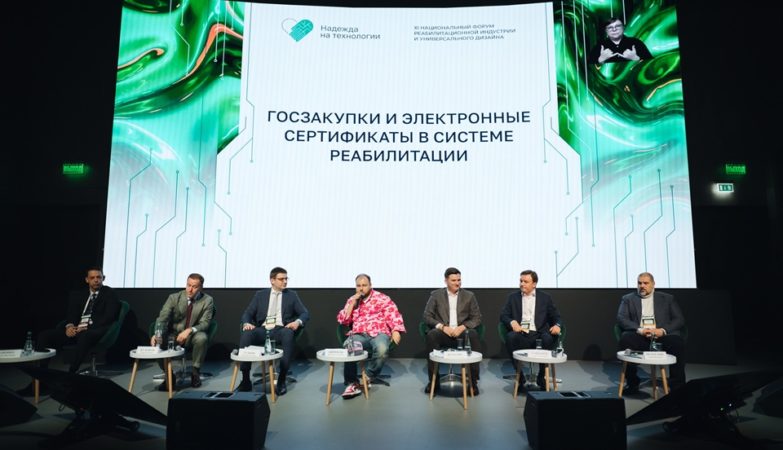 На фотографии запечатлена панельная дискуссия на официальном мероприятии, где на сцене в ряд сидят семеро мужчин-спикеров в костюмах, среди которых выделяется один участник в яркой розовой рубашке с цветочными мотивами. На заднем плане расположен огромный экран с надписью «ГОСЗАКУПКИ И ЭЛЕКТРОННЫЕ СЕРТИФИКАТЫ В СИСТЕМЕ РЕАБИЛИТАЦИИ», логотипом «Надежда на технологии» и окном трансляции с сурдопереводчиком.