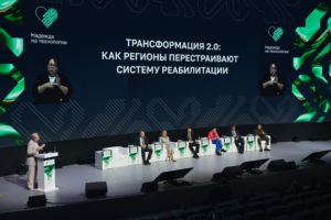 На фотографии показан общий план дискуссионной площадки форума «Надежда на технологии», где спикеры сидят на сцене, а слева за трибуной выступает женщина на фоне огромного экрана с надписью «ТРАНСФОРМАЦИЯ 2.0: КАК РЕГИОНЫ ПЕРЕСТРАИВАЮТ СИСТЕМУ РЕАБИЛИТАЦИИ» и двумя окнами с сурдопереводчиками.
