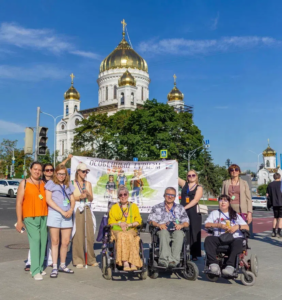 На фотографии запечатлены участники туристической группы, которые улыбаются, и позируют на улице Москвы на фоне величественного Храма Христа Спасителя в солнечный день. Участники экскурсии, среди которых есть люди в инвалидных колясках и с белыми тростями, держат баннер с надписью «Особенный туризм»