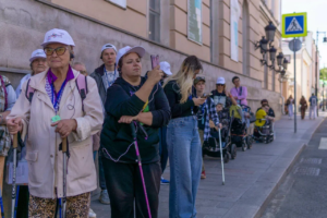 На фотографии запечатлена группа людей в одинаковых белых бейсболках, которые идут по городскому тротуару вдоль исторического здания во время инклюзивной экскурсии. Некоторые участники используют скандинавские и тактильные трости, а женщина на переднем плане фотографирует происходящее на свой смартфон.