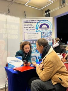 На фотографии запечатлена встреча мужчины и женщины за информационным столом ГУП «Водоканал Санкт-Петербурга» на ярмарке вакансий. Представительница компании показывает что-то в своем смартфоне соискателю, пока они сидят на фоне крупного рекламного баннера с перечнем свободных позиций.